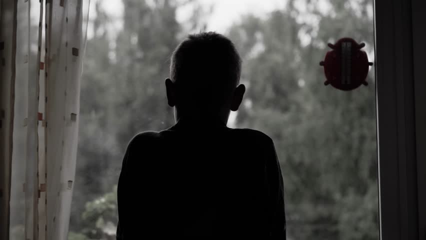 The silhouette of a sad boy who came to the window and looks at the bad weather outside. Very sad footage