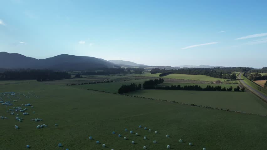 Aerial panoramic drone view of farmland near Christchurch, NZ, with lush green fields, hedgerows, and dramatic mountain ranges in the background under a bright blue sky with scattered clouds.
