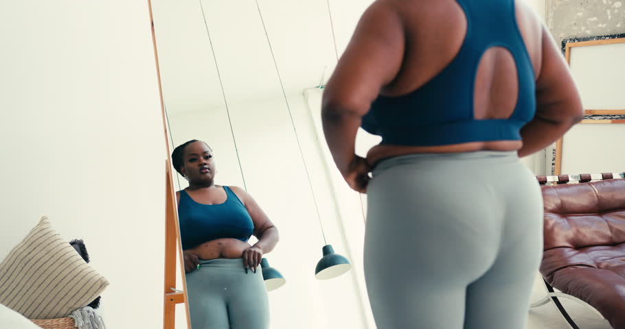 Curvy African American woman in sportswear looks at body in mirror at home. Female pinches excess fat on stomach expressing concern about weight