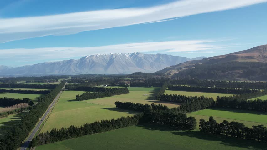 Spanning aerial drone shot near Christchurch, NZ, sweeping across lush farmland and hedgerows to reveal dramatic mountain ranges under a bright blue sky with scattered clouds.
