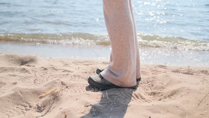 A woman with long blonde hair in an ethnic patterned jacket walks the sandy shore toward a man and embraces him tightly. The calm sea and clear spring sky emphasize their romantic warmth.