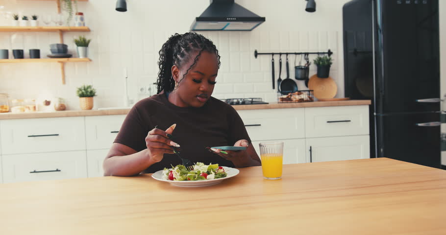 Overweight black woman sitting at kitchen table eats fresh salad. Female looks at phone having healthy meal and staying connected to online world