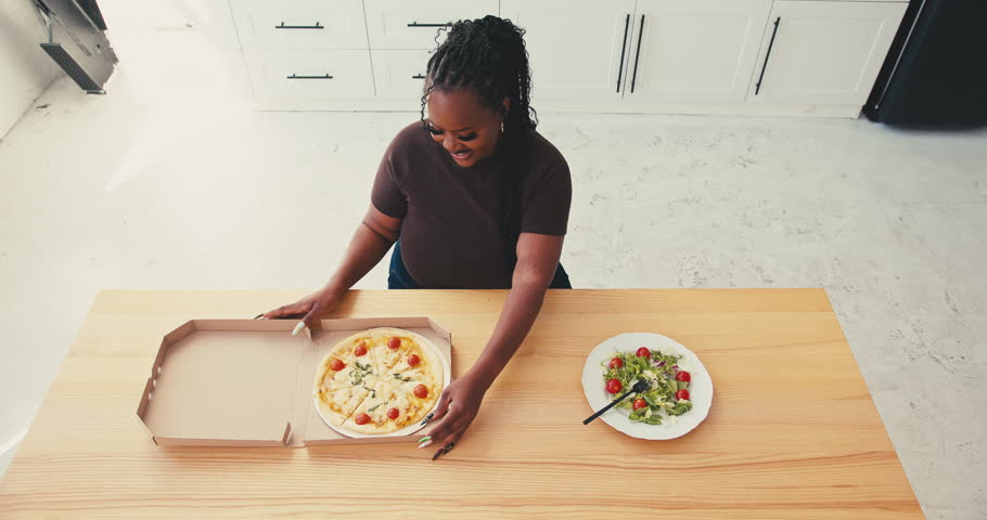 Plus size black woman finally makes choice after much deliberation between healthy salad in favor of pizza. Smile appears on lady face immediately