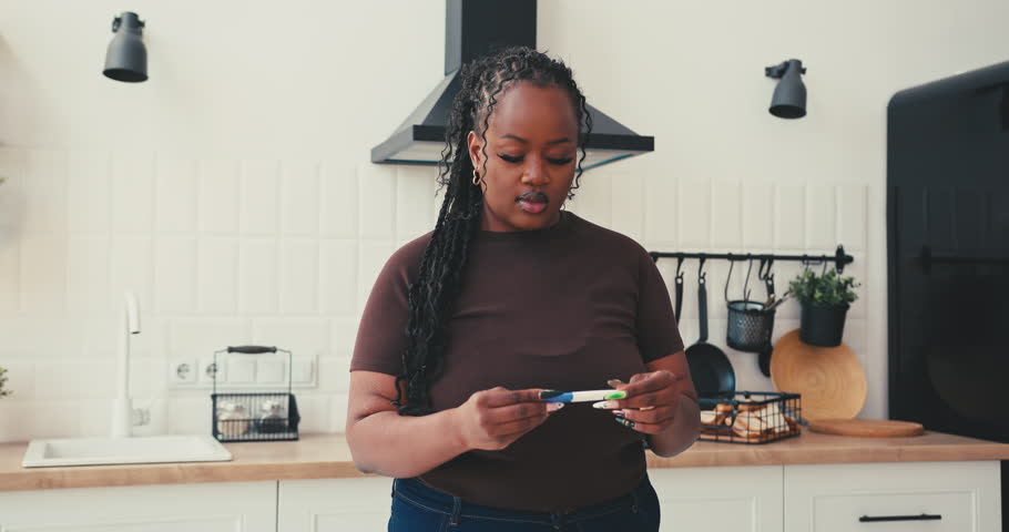 Plus size black woman holds pregnancy test anxiously examining result. Female shows extremely happy emotions after seeing positive picture in kitchen