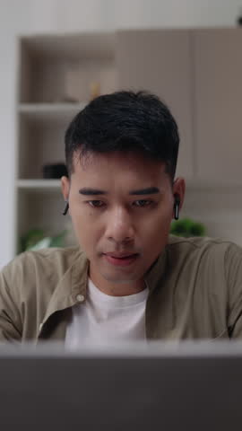 Asian man smiling at camera while working on laptop in home office. Concept of remote work, online communication, friendly professional, digital lifestyle, and positive freelance routine.