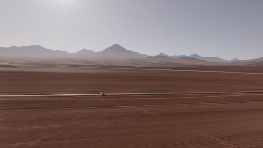 Aerial shot of a car driving through a scenic road in the Atacama highlands surrounded by mountains and volcanoes under the summer sun in one of the most remote landscapes in Chile