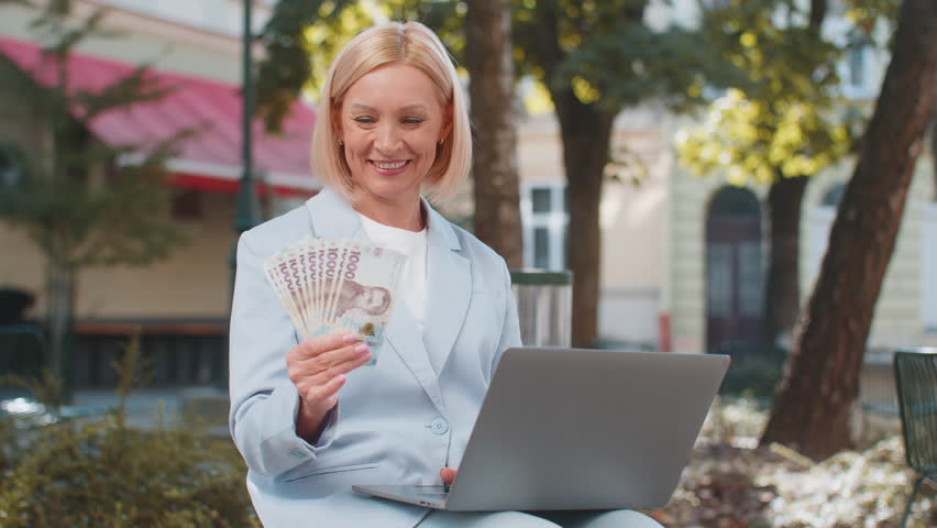 Happy mature business woman investor counting Ukrainian hryvnias UAH banknotes on city street. Smiling entrepreneur lady looking at laptop holding money and rejoices at good earnings. Success lottery