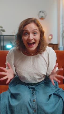 Happy young woman watching TV shouting, celebrating success, winning, goal achievement good news, lottery luck, victory sitting on sofa in living room. Girl showing double thumbs up at home. Vertical