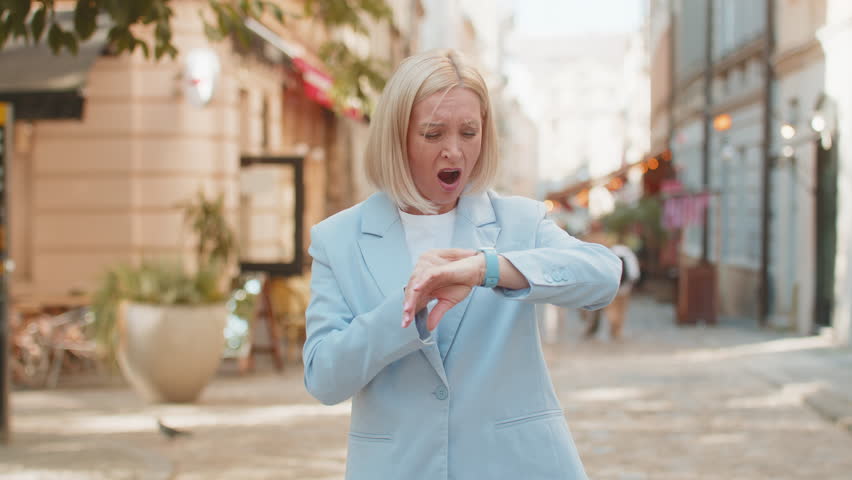 Shocked mature woman businesswoman in hurry checking time on smartwatch walking on city street. Medium shot of Caucasian middle-aged female entrepreneur late for business meeting walk fast outside.