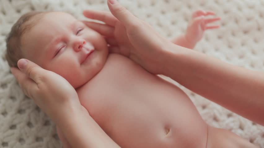Close-up of a smiling baby holding mother hands while lying on a soft blanket indoors, showing joy, tenderness, bonding, and loving family connection.