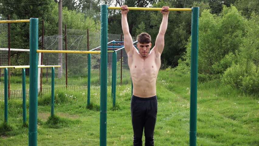 Strong guy training abdominal muscles on horizontal bar at sports ground. Young sportsman exercising on pull-up bar outdoor. Sporty athlete working out at nature. Concept of sportive lifestyle