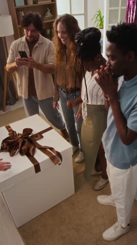 Vertical high angle view of intrigued multiethnic friends waiting for future parents to open lid of large box with blue balloons, clapping and feeling overjoyed after revealing babys gender
