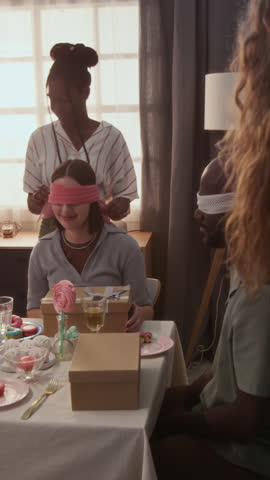 Vertical view of blindfolded young spouses opening boxes, taking baby shoes out of them and trying to guess when having fun game at holiday table during home gender reveal party