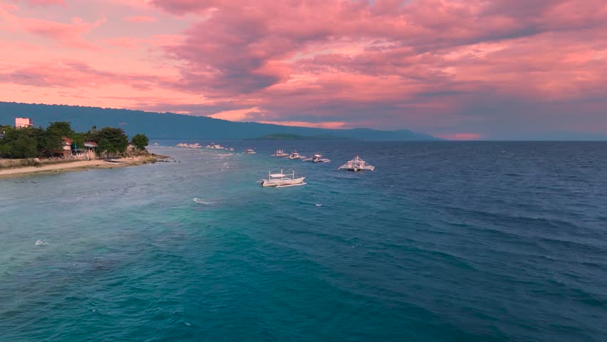 Aerial view of traditional Filipino outrigger boats floating on the ocean near a coastline with houses, under a vibrant pink and orange sunset sky, Boracay, Philippines.