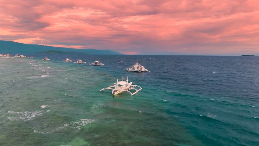 Aerial view of traditional Philippine outrigger boats floating on ocean water under a pink and orange sunset sky, Boracay, Philippines.