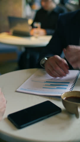 Close up view of two businessmen shaking hands over printed financial report at cafe table during coffee meeting. Vertical format footage