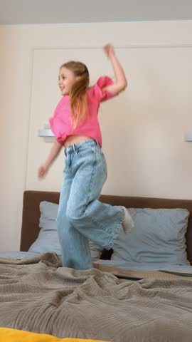 Energetic young girl jumping with pure joy on soft bed, radiating childhood happiness through playful movement in cozy bedroom setting vertical video