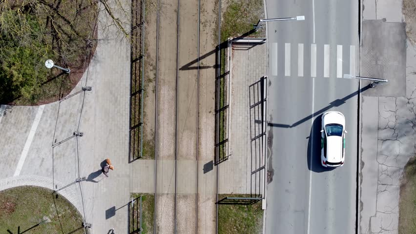 Aerial view of a street intersection with tram tracks, two moving cars, a pedestrian with an orange bag, crosswalks, and streetlights.