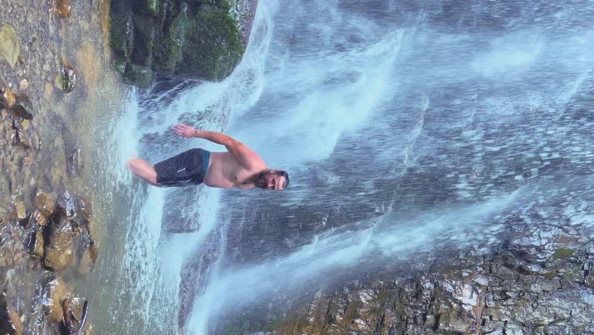 A young attractive traveler stands under the stream of a mountain waterfall. vertical video