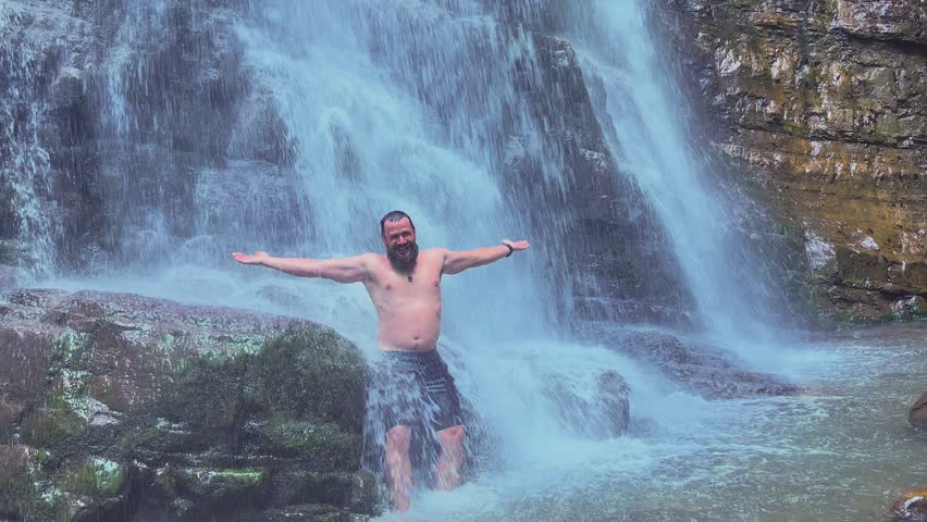 A young attractive traveler stands under the stream of a mountain waterfall. 