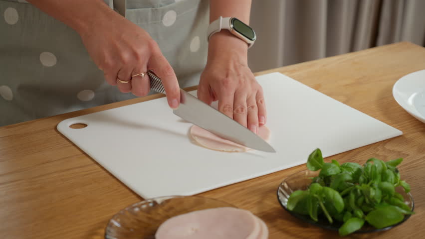 A woman slicing cooked ham into small pieces on a cutting board. The process of preparing meat ingredients for a salad, pizza, omelette, or sandwiches for a tasty homemade meal.