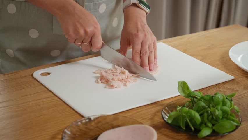 Chopping cooked deli meat into cubes for a delicious breakfast. A woman prepares ingredients for a homemade omelette, frittata, or scrambled eggs with fresh ham and herbs.