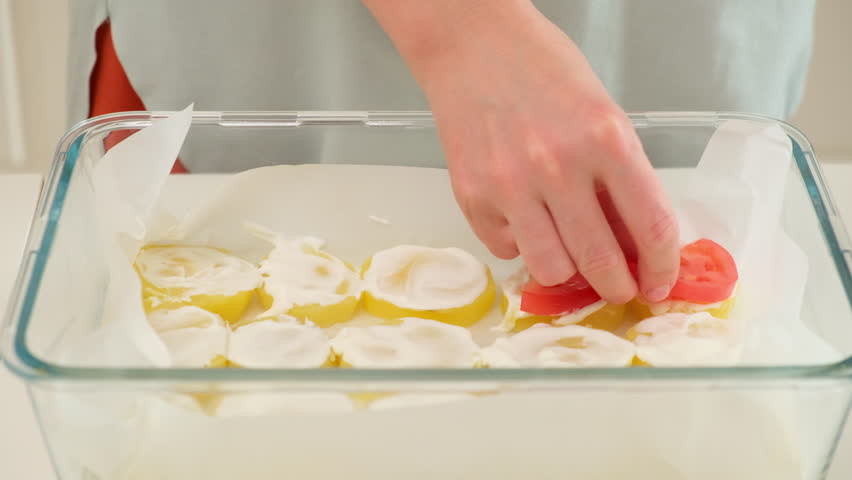 Close-up of a hand placing fresh tomato slices over baked potato rounds topped with sour cream in a glass baking dish.