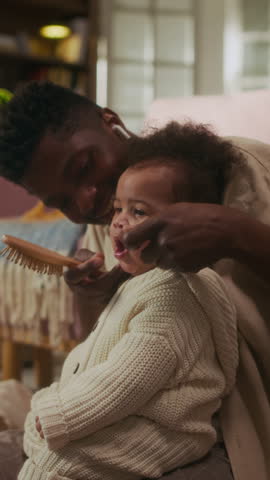 Vertical side view shot of adorable baby girl yawning while sitting in fathers lap with loving African American father brushing her hair
