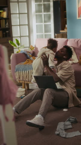 Vertical full length shot of young African American father playing with baby girl while sitting on floor and using laptop trying to work from home
