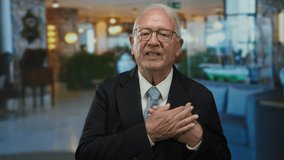 Senior man in a business suit standing indoors at a hotel, gesturing with hands on heart, conveying emotion and professionalism. - Powered by Shutterstock - Get 15% off with code: PIKWIZARD15