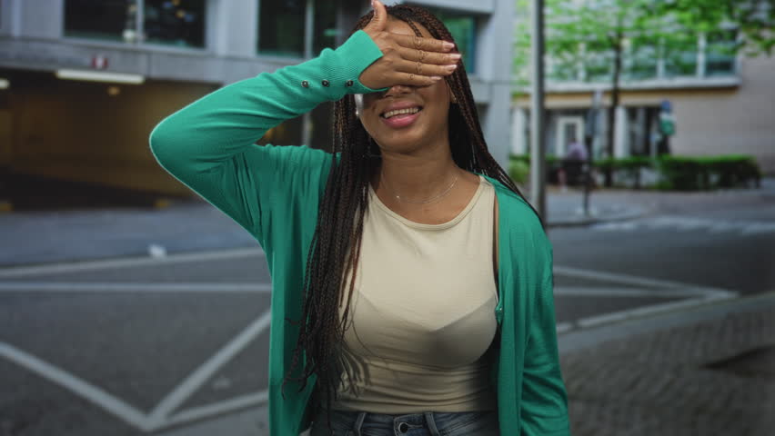 Woman in green cardigan covering eyes with hand and smiling on urban street near building crosswalk; shy joy.