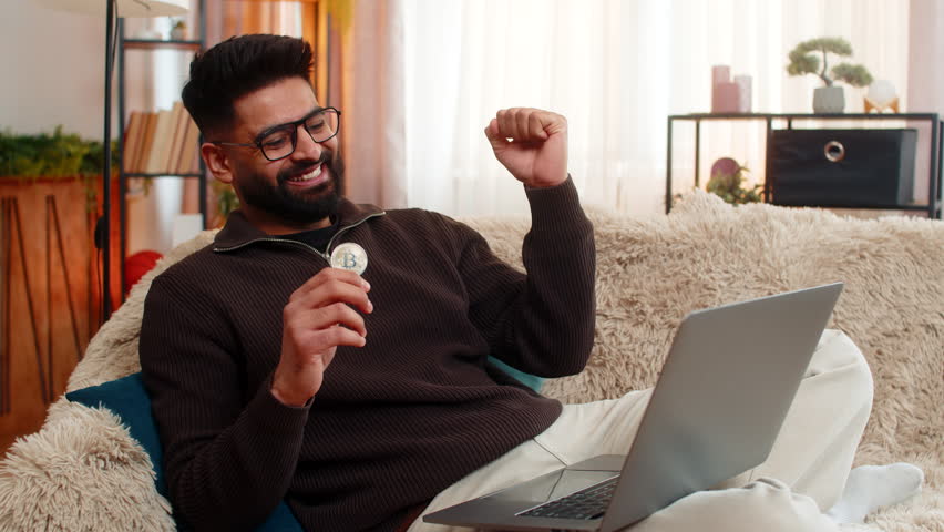 Indian man at home watches Bitcoin BTC chart rise on laptop, smiling while holding coin with confident hand. Arabian guy on sofa shows excitement joy, celebrating crypto success with raised gesture. - Powered by Shutterstock - Get 15% off with code: PIKWIZARD15