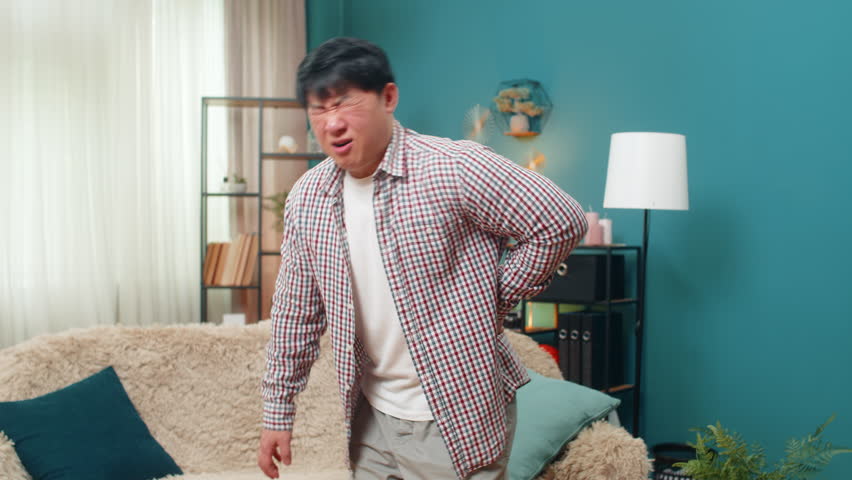 Asian man on home sofa grimaces, lowering body carefully, clutching back as sharp pain strikes. Chinese guy shows intense discomfort, tense face stiff posture revealing spinal strain physical stress