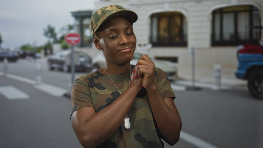 Woman in camouflage uniform clasps hands and dogtags while wearing a military hat on street near historic building; pride duty.