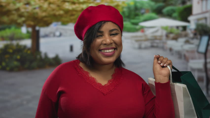 Woman smiling in red clothing holds shopping bags on a city terrace with outdoor seating and greenery, showcasing youthful joy and urban shopping experience.