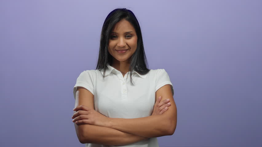 Woman smiling with arms crossed, wearing white shirt, standing against a purple background, exuding confidence and happiness in an isolated studio setting.