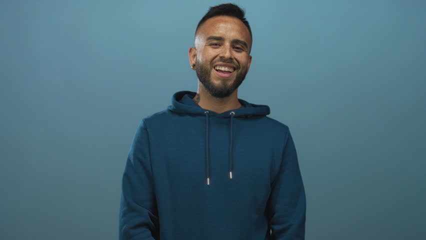 Man smiling showing teeth and beard in teal studio wearing blue hoodie, relaxed pose; joy warmth openness.