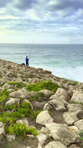 Unrecognizable male adult stands on jagged rocks by ocean, casts fishing line into water as waves crash nearby. Dramatic sky, with clouds, serene fishing atmosphere, cloudy dramatic sky over clear
