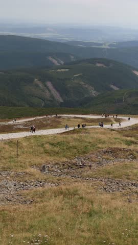 Vertical screen mountain trail winds through green slopes as groups of hikers walk along the stony path surrounded by vast valleys and forested hills creating a sense of freedom and adventure