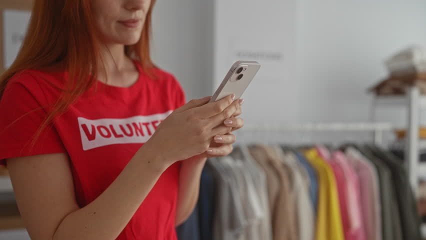 Woman in red shirt holds phone in charity center with clothing rack emphasizing volunteer activity and community support indoors.
