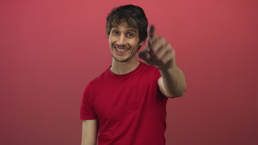 Young man wearing red shirt smiling and making heart gesture with hands in studio against red backdrop; love.