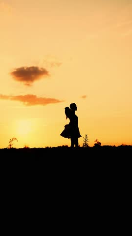 Silhouette of a young happy girl in a dress spinning around herself with her arms outstretched at sunset in a field. The woman enjoys life and a beautiful summer sunset. Vertical video.
