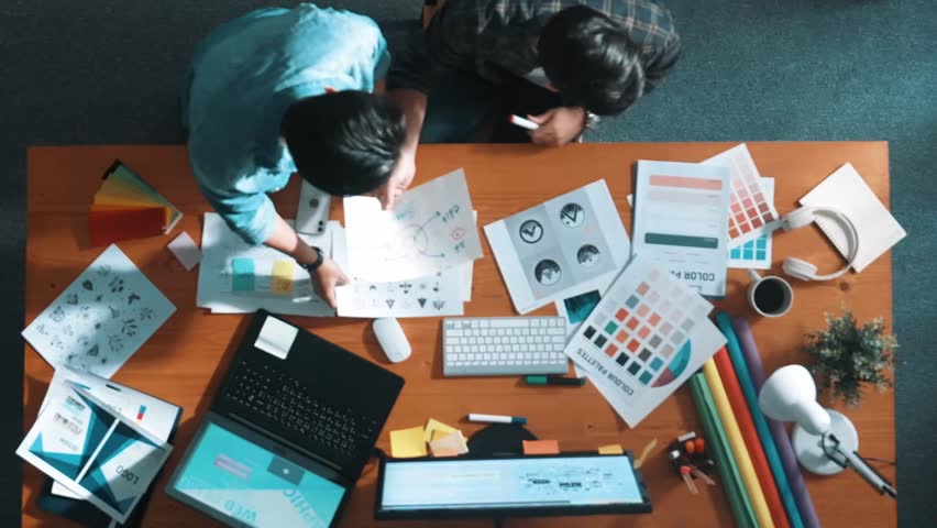 Top down aerial view of project manager looking at screen while designer writing idea. Smart business team explain idea of logo design while designer choose theme while looking at palette. Symposium.