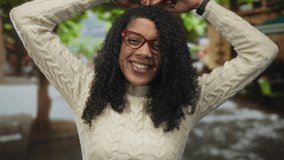 Woman touches hair with hands in park under green trees while smiling broadly and wearing red glasses and a cream sweater; happiness. - Powered by Shutterstock - Get 15% off with code: PIKWIZARD15