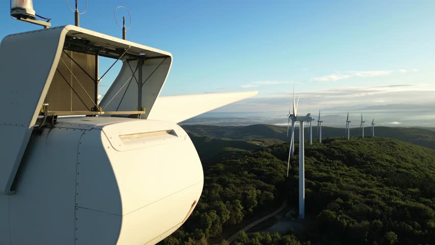 A close-up view of a wind turbine on a hill at sunrise, blades spinning among trees, captured in a scenic aerial shot showcasing renewable energy in a natural setting.