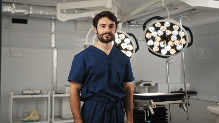 A smiling surgeon with crossed arms stands in an operating room, looking directly at the camera. A male doctor, possibly of Mediterranean descent, prepares for surgery in a modern operating room - Powered by Shutterstock - Get 15% off with code: PIKWIZARD15