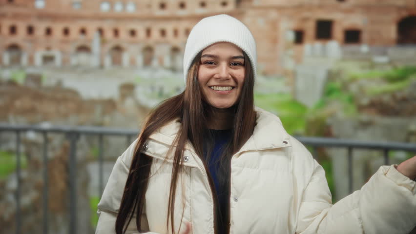 Young woman joyfully mimicking guitar playing in front of ancient roman ruins wearing a white jacket and hat in an outdoor street setting with a warm smile and long hair flowing.