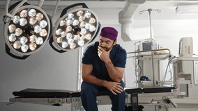 A male surgeon sits pensively in the operating room, resting his face in his hand, a purple cap on his head. A concerned doctor in a surgical setting, illuminated by bright lights, expressing worry. - Powered by Shutterstock - Get 15% off with code: PIKWIZARD15