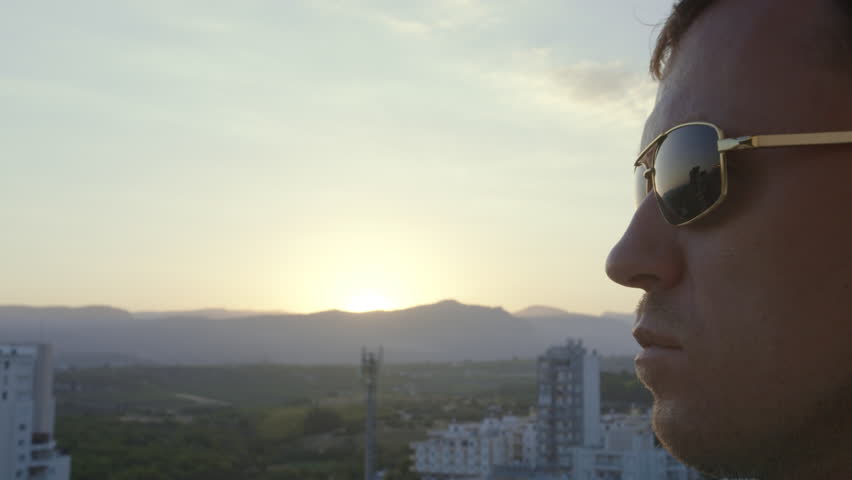 Man wearing sunglasses, takes off his sunglasses and watches the sun go down behind the mountains, camera movement