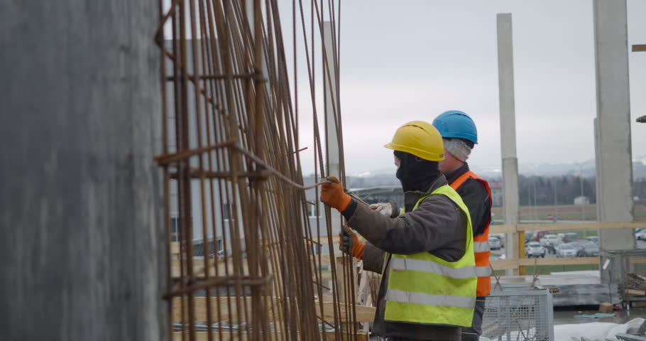 Cracow, Poland – October 10, 2019: Two steel fixers in hard hats and safety vests work together to build a reinforcement cage. They are manually tying steel rebar to create the structural skeleton.